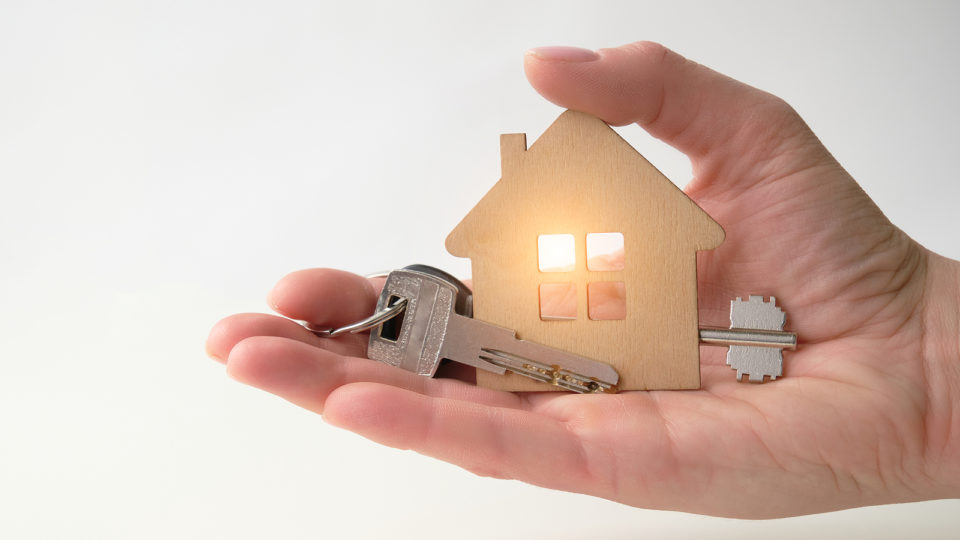 A Womans Hand Holds A Model Of A Wooden House And Keys. The Conc